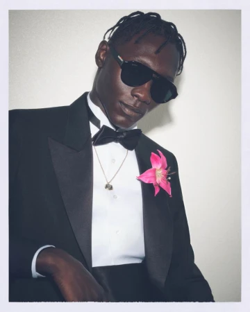 Black man wearing dark sunglasses, tuxedo, and a pink flower on the lapel against a plain background.