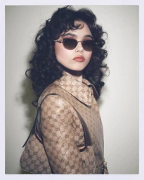 Model with dark curly hair wearing monogram patterned jacket and sunglasses against plain background