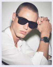 Close-up portrait of a male model wearing black sunglasses and a white shirt against a plain background.