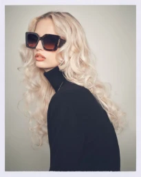 Blonde woman wearing dark oversized sunglasses and a black turtleneck against a neutral studio background
