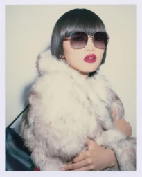 Model wearing dark oversized sunglasses and a textured fur coat against a plain white studio background