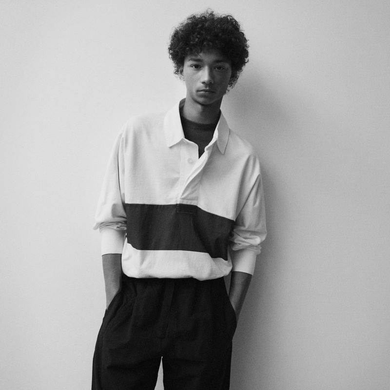 Monochrome portrait of a young adult male with curly hair wearing a long-sleeve collared shirt and dark pants posing against a plain wall.
