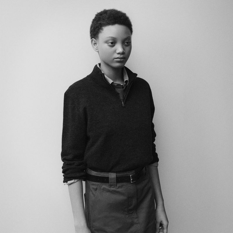 Model wearing dark quarter-zip sweater and belted skirt standing against plain studio background