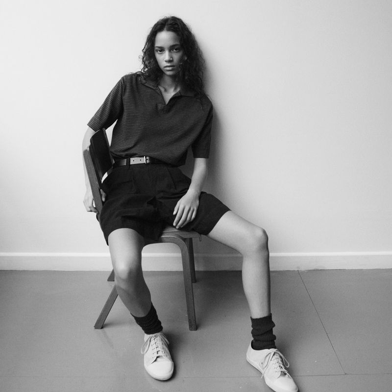 Model wearing a collared top and shorts sitting on a wooden chair against a plain white studio background.