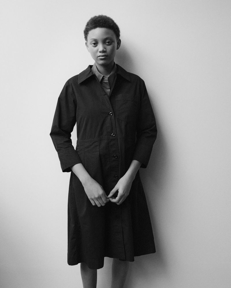 Monochrome portrait of a young Black woman in a button-down dress, standing against a neutral wall with soft, diffused lighting.