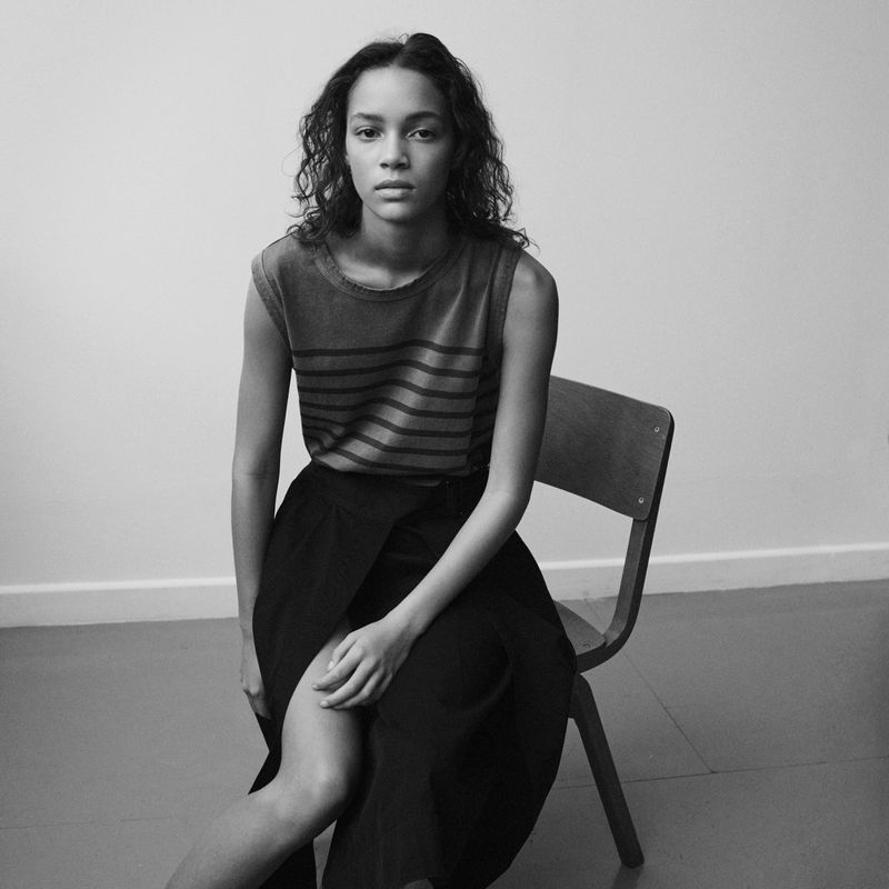 Black-and-white shot of a curly-haired model in a striped top and black skirt, seated on a wooden chair against a plain background.