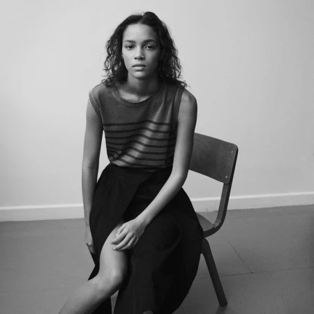 Black-and-white shot of a curly-haired model in a striped top and black skirt, seated on a wooden chair against a plain background.