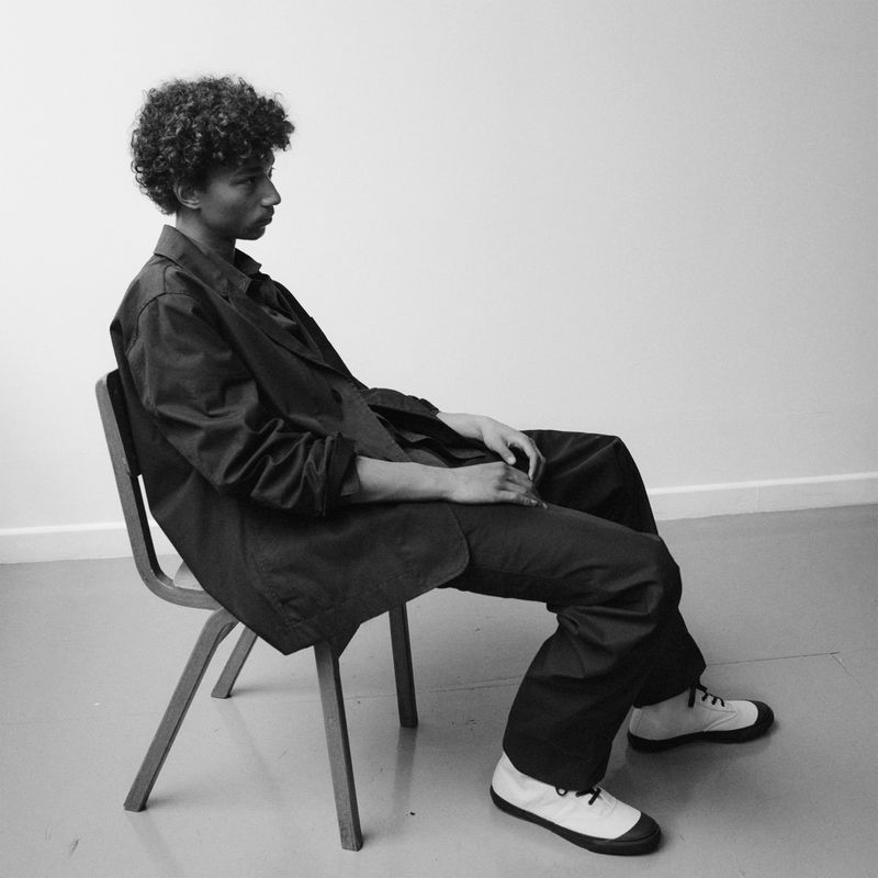 Monochrome portrait of a Black male model with curly hair in a dark suit sitting on a wooden chair against a white wall backdrop.