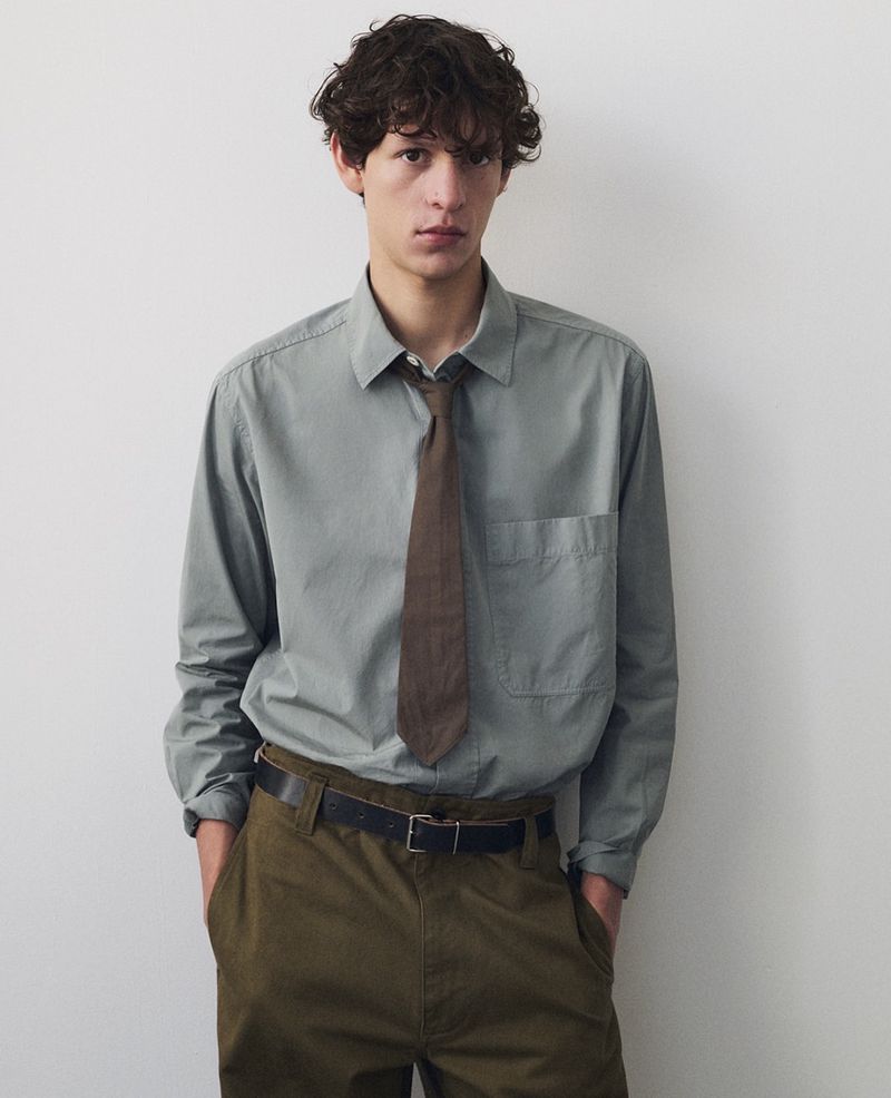 Portrait of a young adult man with curly hair wearing an olive-green long-sleeved shirt and tie, standing against a neutral background.