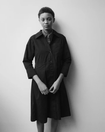 Black and white portrait of a model wearing a dark buttoned coat against a plain light background for MHL by Margaret Howell.