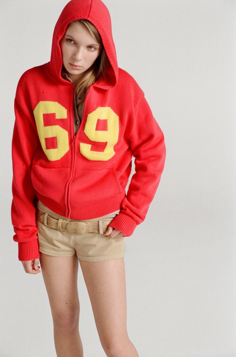 A fair-skinned young woman stands indoors wearing a red hooded sweatshirt with yellow '69' numbers and khaki shorts against a neutral backdrop.