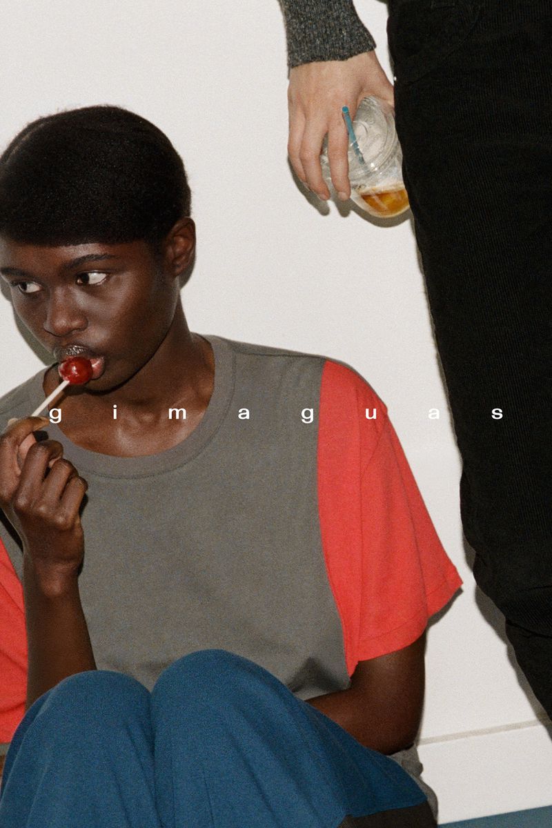 Young Black woman with lollipop poses indoors near another person holding a beverage, in a studio environment with modern fashion.