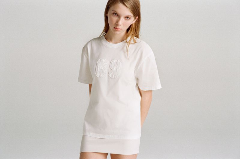 A woman in her early twenties poses in a white t-shirt and skirt against a plain, neutral background, showcasing a minimalist style.