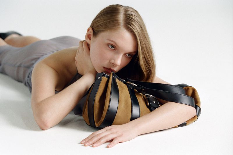 A fair-skinned woman lying down in a gray dress with a brown-and-black purse in a studio setting with a white background and neutral tones.