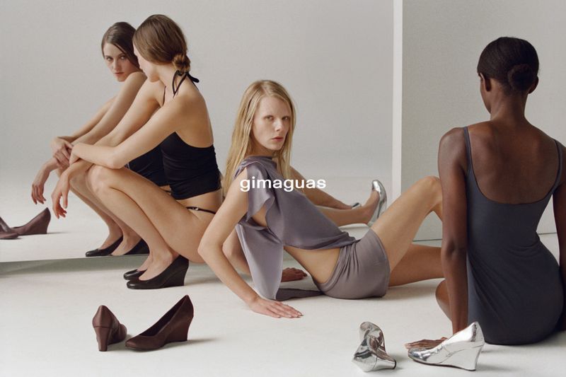 Four women pose in a studio setting, showcasing wedge heels in black and silver, with a clean white backdrop and minimalist styling.