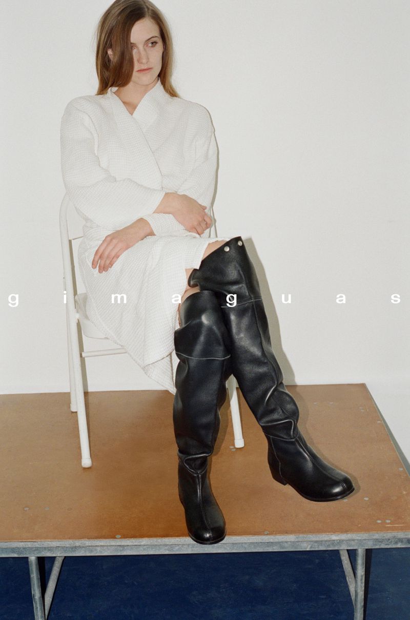 A woman in a white robe sits on a chair wearing black leather thigh-high boots against a white background in a fashion studio setting.