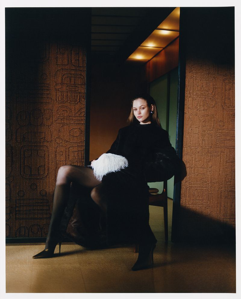Elegant adult woman in black dress, tights, and heels, seated in chair with a white feather accessory against textured wall.
