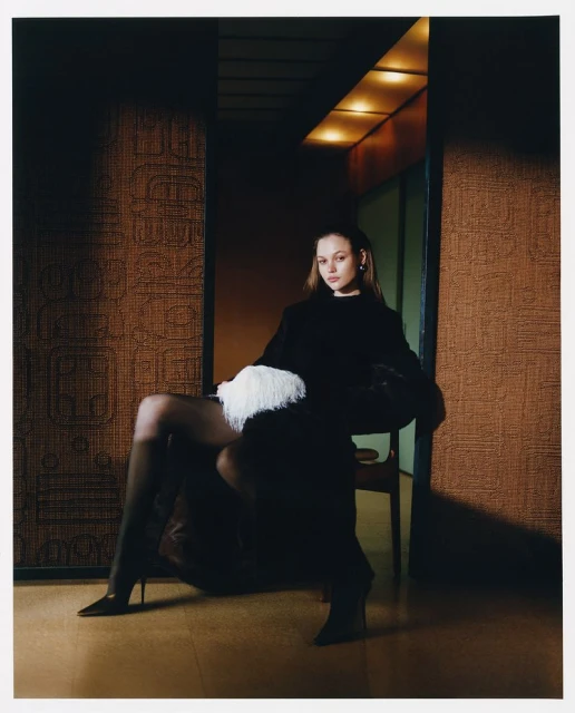 Elegant adult woman in black dress, tights, and heels, seated in chair with a white feather accessory against textured wall.