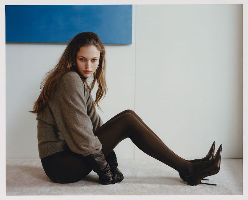 Indoor fashion portrait of an adult woman with long brown hair wearing a grey sweater, black tights, and high heels sitting on the floor.