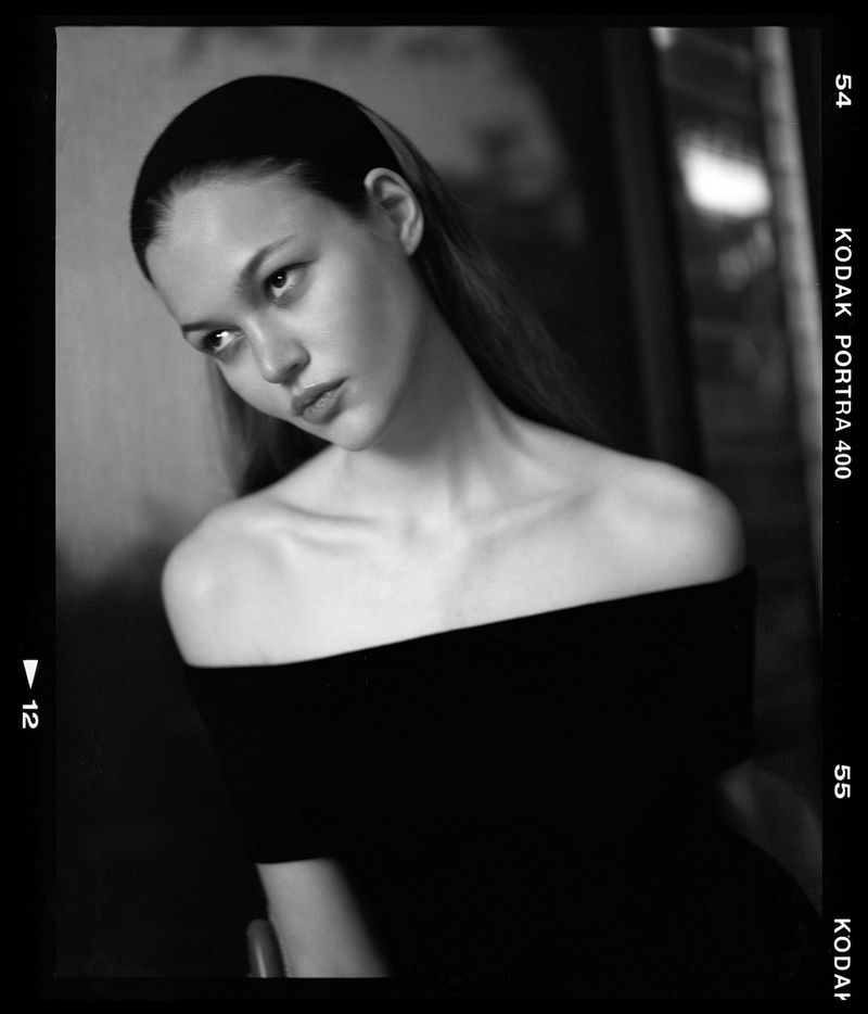 Monochrome portrait of a young adult woman with bare shoulders wearing a headband, looking off-camera with soft lighting and film grain.