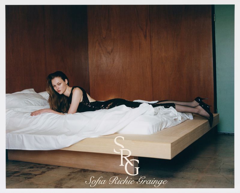 Woman in black sequin dress lying on white bed in warm interior, wearing high heels, medium shot, indoor fashion portrait.