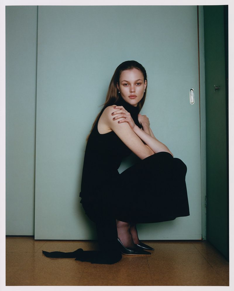 A young woman poses crouching in a black dress and black heels against a teal sliding door in a studio setting.