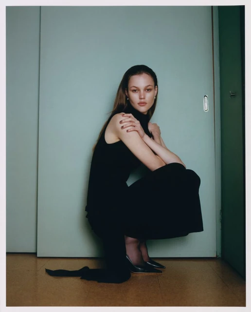 A young woman poses crouching in a black dress and black heels against a teal sliding door in a studio setting.