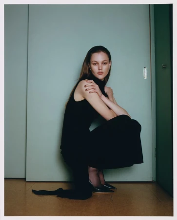 A young woman poses crouching in a black dress and black heels against a teal sliding door in a studio setting.