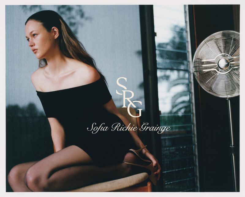 Portrait of a young woman with long brown hair sitting in a short black dress in front of a window with a metal fan.