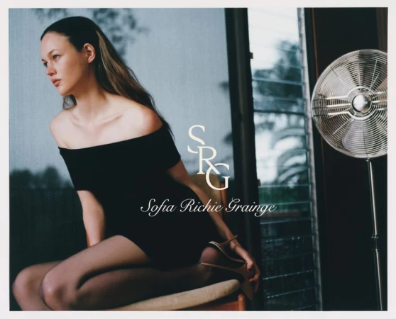 Portrait of a young woman with long brown hair sitting in a short black dress in front of a window with a metal fan.