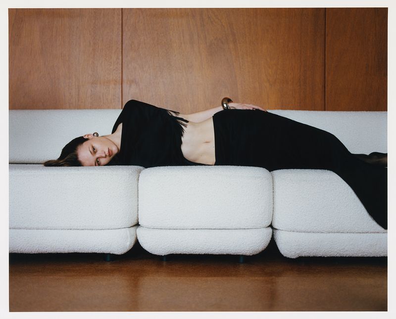 Indoor shot of a woman in black laying on a white couch, with a wooden wall background, in a lifestyle or fashion setting. 