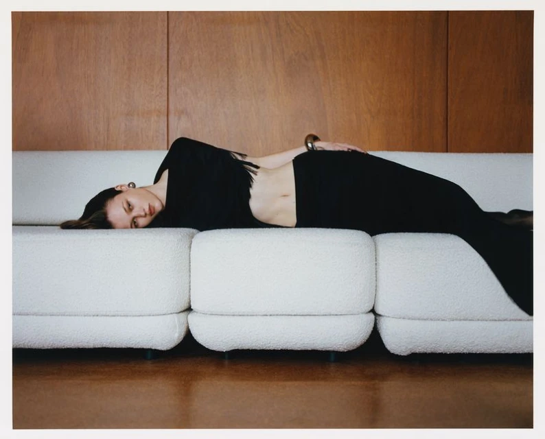 Indoor shot of a woman in black laying on a white couch, with a wooden wall background, in a lifestyle or fashion setting. 