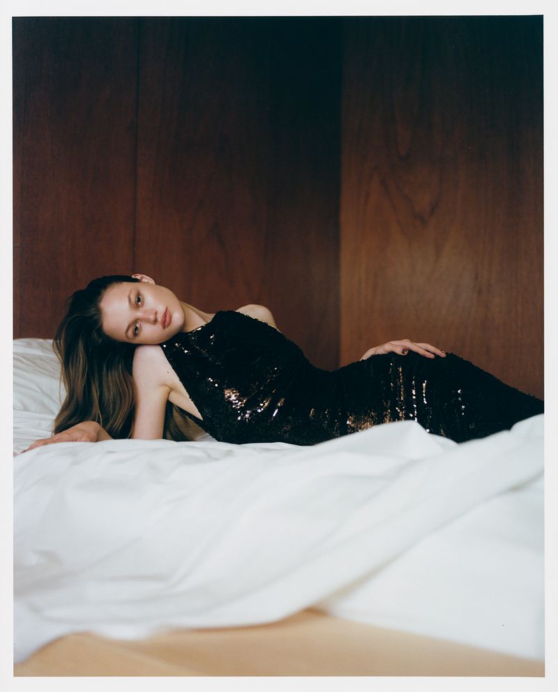 Fashion shot of a woman in a black sequin dress lying on a white bed against a wood paneled wall in a warm, low-key lighting setting.