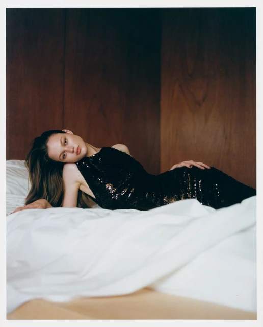 Fashion shot of a woman in a black sequin dress lying on a white bed against a wood paneled wall in a warm, low-key lighting setting.