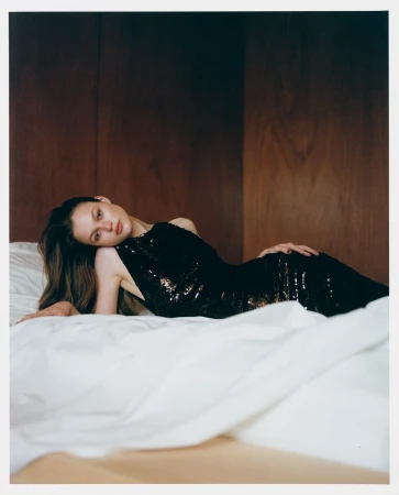 Fashion shot of a woman in a black sequin dress lying on a white bed against a wood paneled wall in a warm, low-key lighting setting.