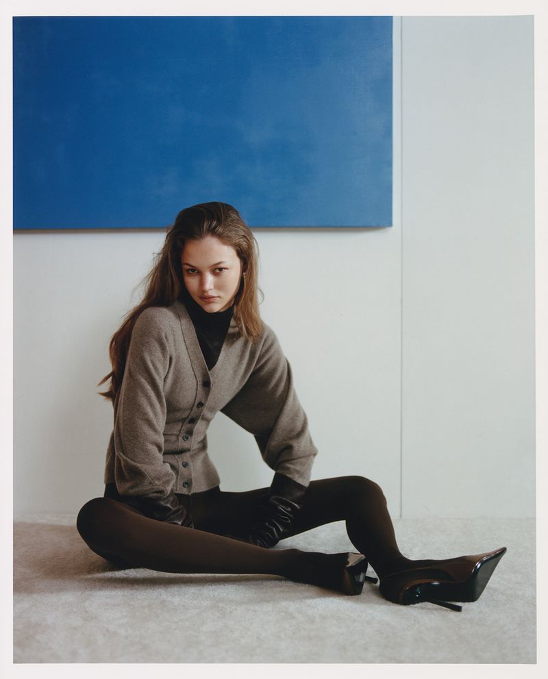 A woman sits on the floor in black tights and high heels with a grey cardigan and a blue square painting on the wall behind her.