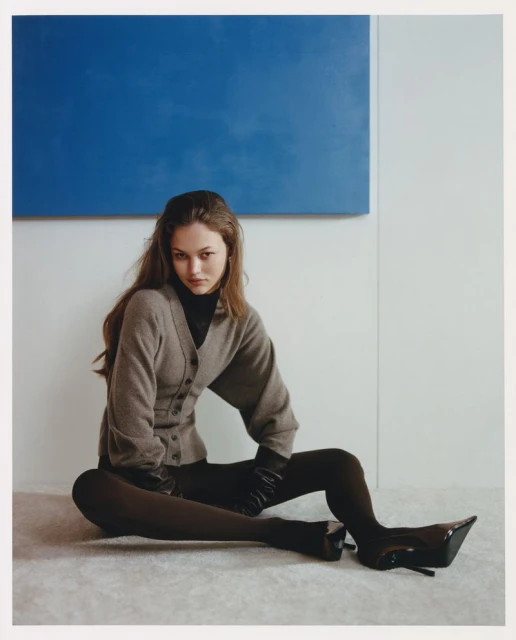 A woman sits on the floor in black tights and high heels with a grey cardigan and a blue square painting on the wall behind her.
