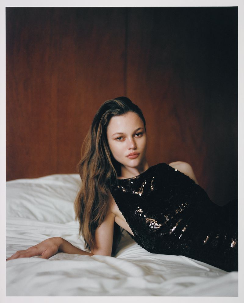 A woman with long brown hair wearing a black sequined dress lies on a white bed in a studio, posing for a portrait in soft lighting.