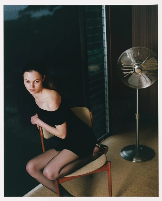 Young woman in black dress poses seated in a room with dark walls, an upright fan, and sunlight streaming through the window.