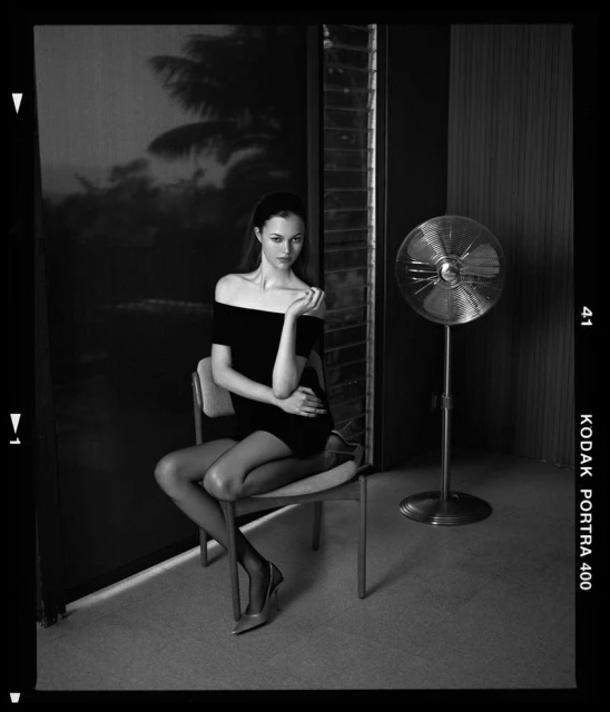Black and white portrait of a young woman in a short dress and heels sitting on a chair in a room with a fan and window.
