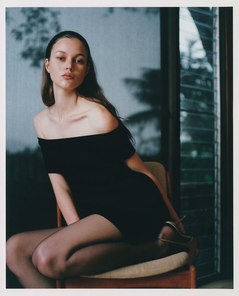 Elegant young woman posing on a chair in a black off-the-shoulder dress and sheer stockings with window and greenery in the background.