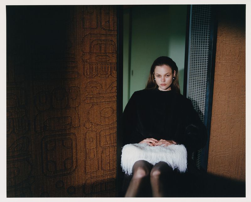 A young woman with long hair sits in front of a patterned wall, wearing a black top and a white, fringed skirt in a dimly lit room.