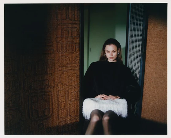 A young woman with long hair sits in front of a patterned wall, wearing a black top and a white, fringed skirt in a dimly lit room.