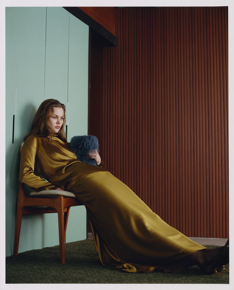 Young woman in a golden gown poses in a room with a green cabinet, dark brown ridged wall, and green carpet. Lifestyle fashion photography.