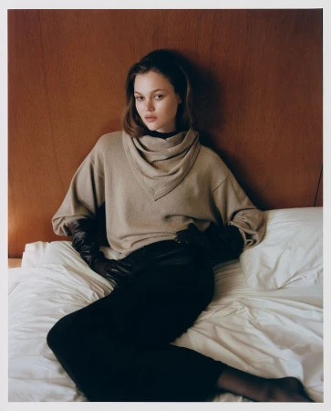 Model wearing a beige cowl-neck sweater and dark pants on a bed against wood paneling
