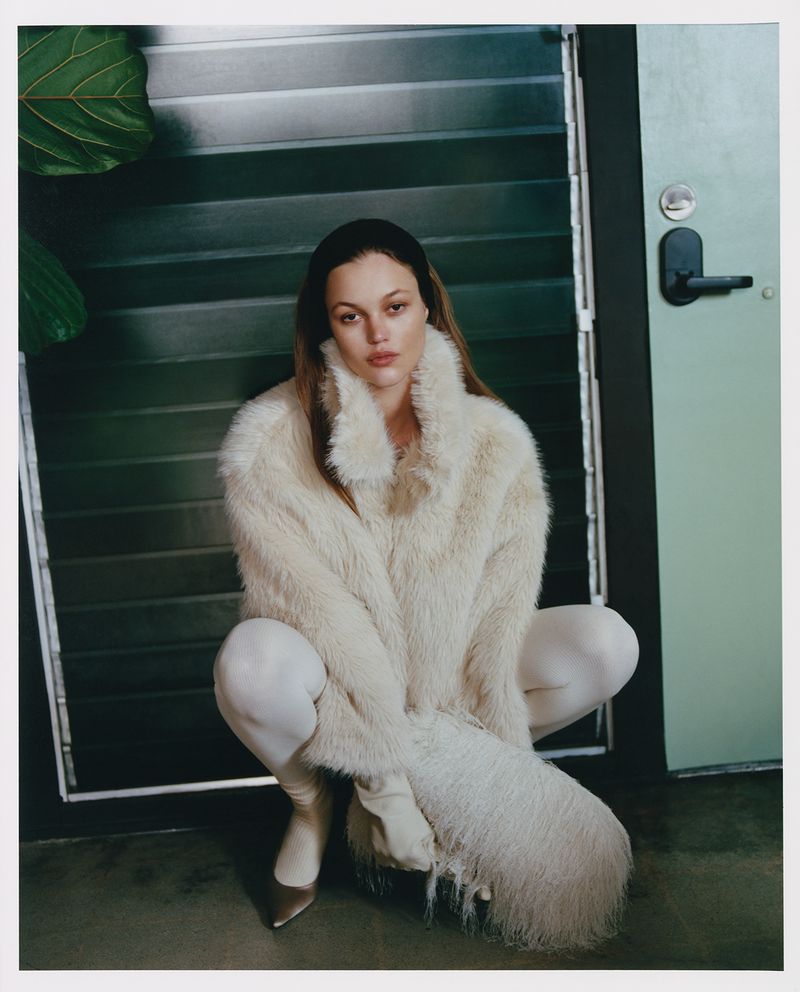 A woman with long brown hair and a headband poses while squatting. She is wearing white leggings, brown heels, and a cream-colored fur coat.