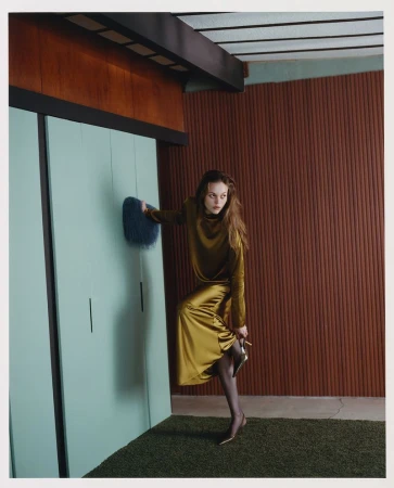 A stylish young woman in a gold dress and black tights poses indoors by a teal wall with a retro shag rug and brown wood paneling.