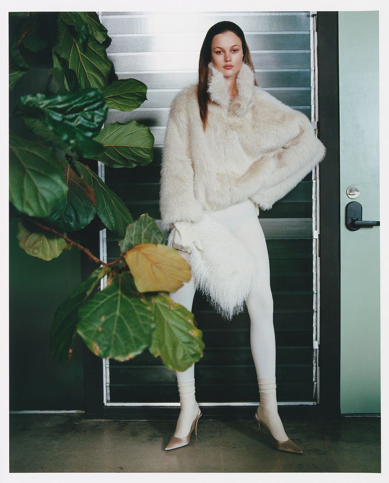 A model poses indoors wearing a cream faux-fur jacket, white pants, and heels with a large green plant to the side, fashion shot.