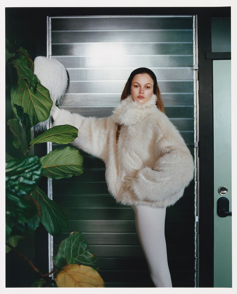 Young woman posing in a white faux fur coat next to a large plant in an indoor setting with light coming through slatted blinds.