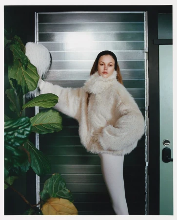 Young woman posing in a white faux fur coat next to a large plant in an indoor setting with light coming through slatted blinds.